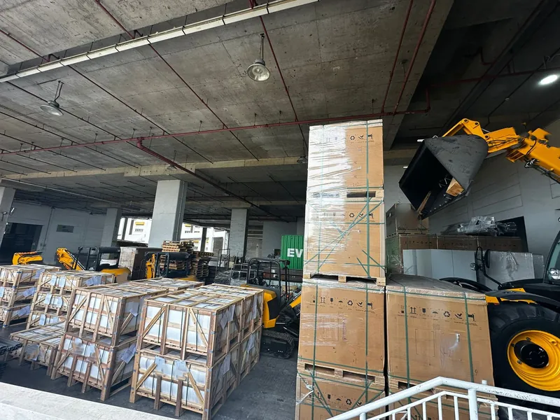 Crated and palletized cargo stacked at a covered loading bay beside a telehandler